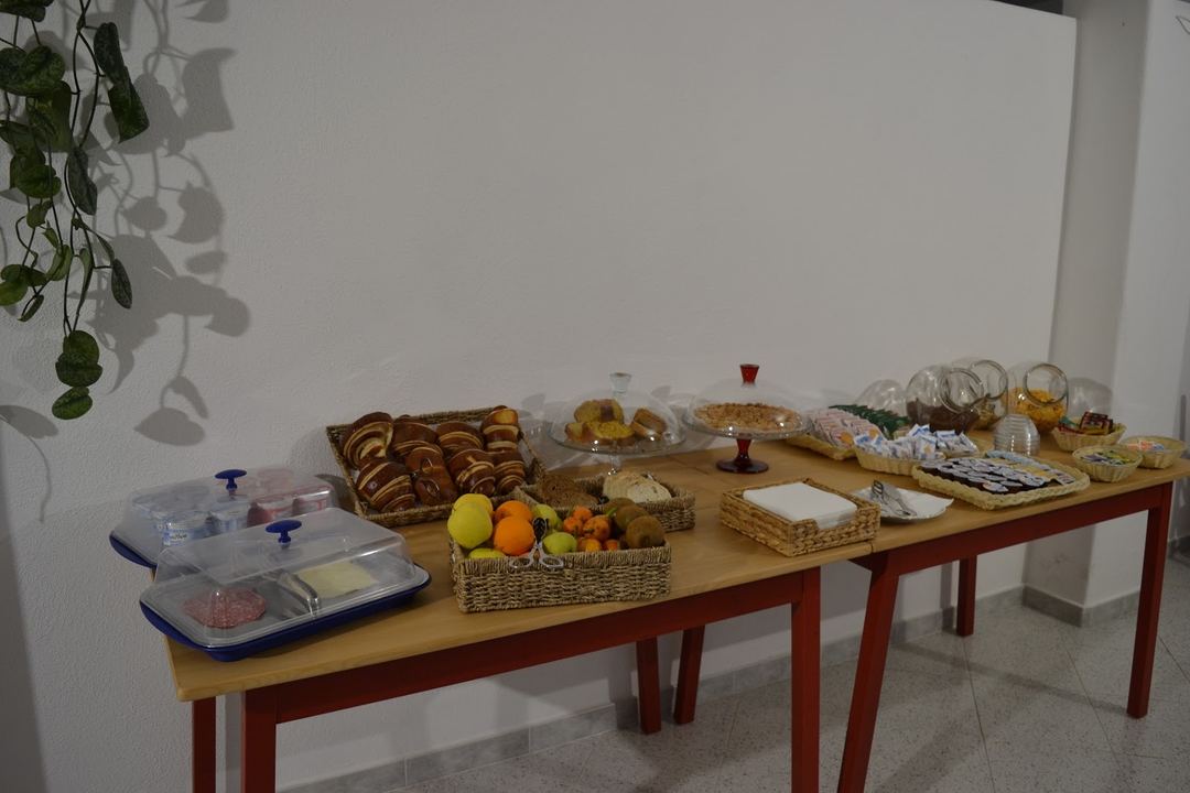 Breakfast buffet display featuring fresh fruits, pastries, breads, jams, oils, and beverages arranged on a wooden table with red legs.