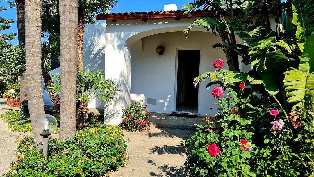 A charming white bungalow with curved architecture surrounded by vibrant pink flowers and palm trees at Cocos Park Ischia.