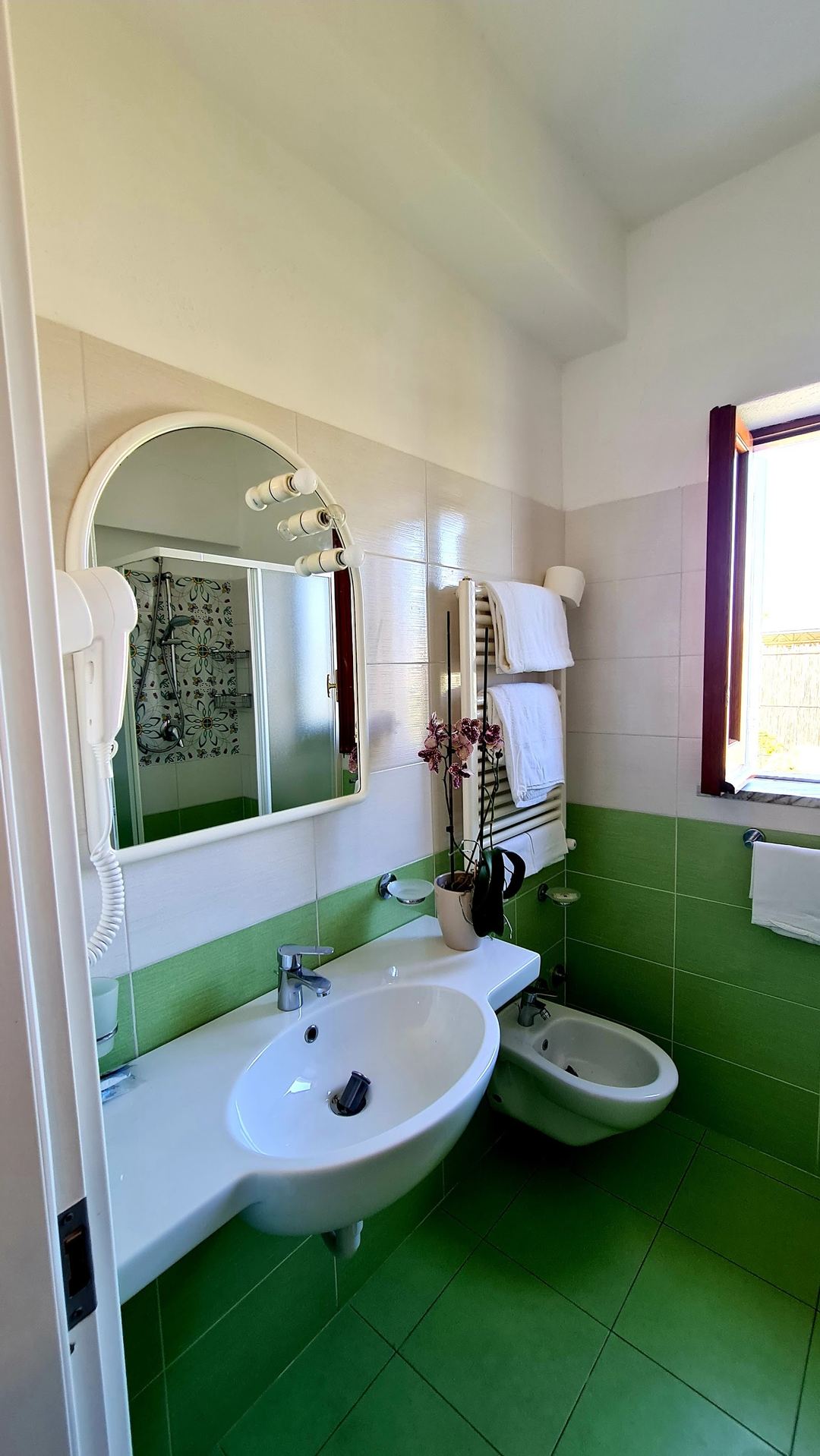 A modern bathroom with bright green tile flooring, white fixtures, illuminated mirror with sconces, and towel rails.